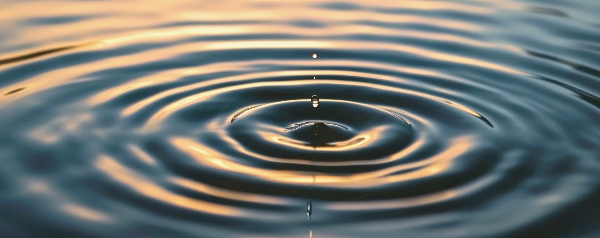 A single water droplet meeting a still, deep blue surface, sending calm ripples outward — a visual metaphor for small changes creating lasting momentum.
