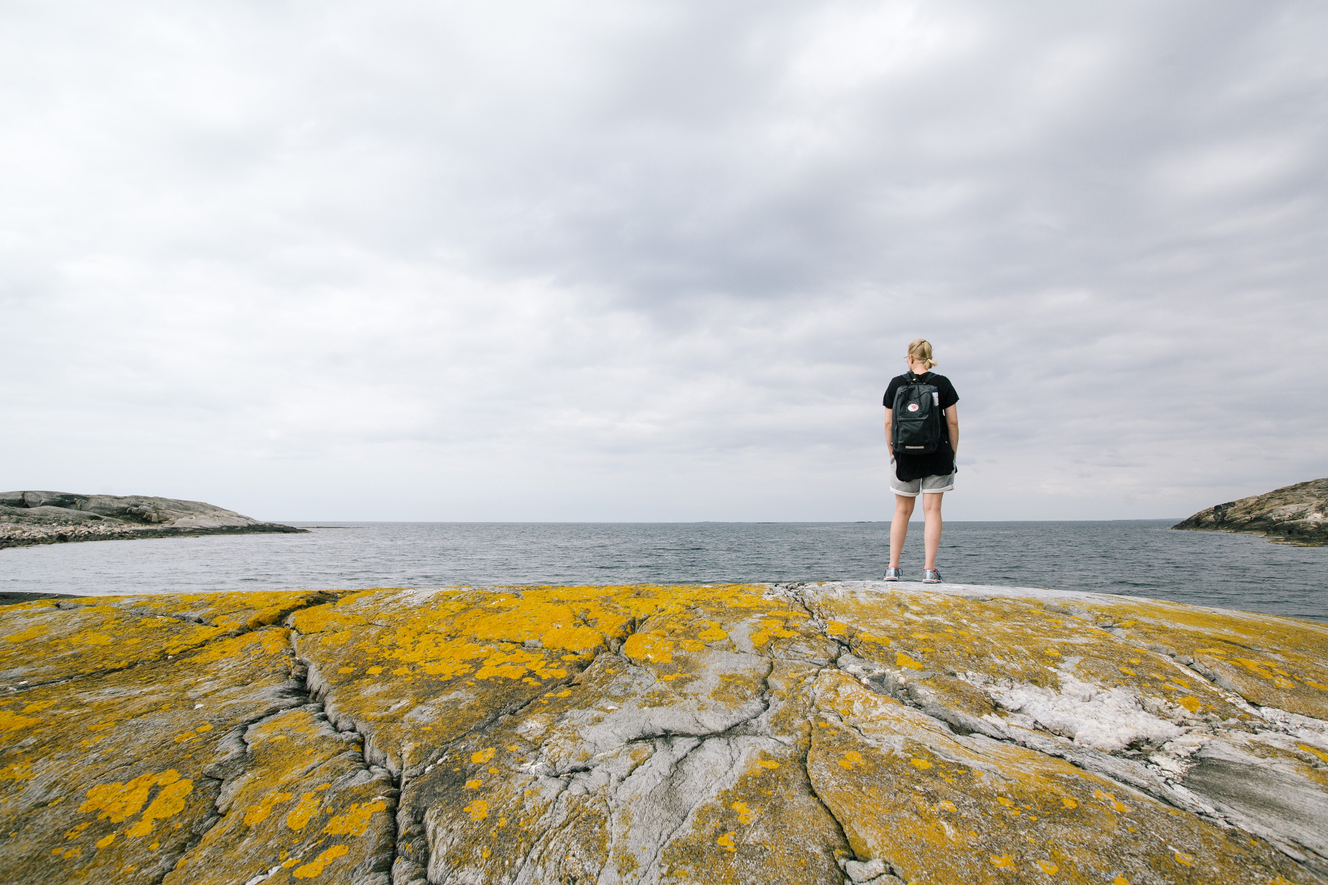 A woman with a backpack standing on moss-covered rocks by the sea under a cloudy sky