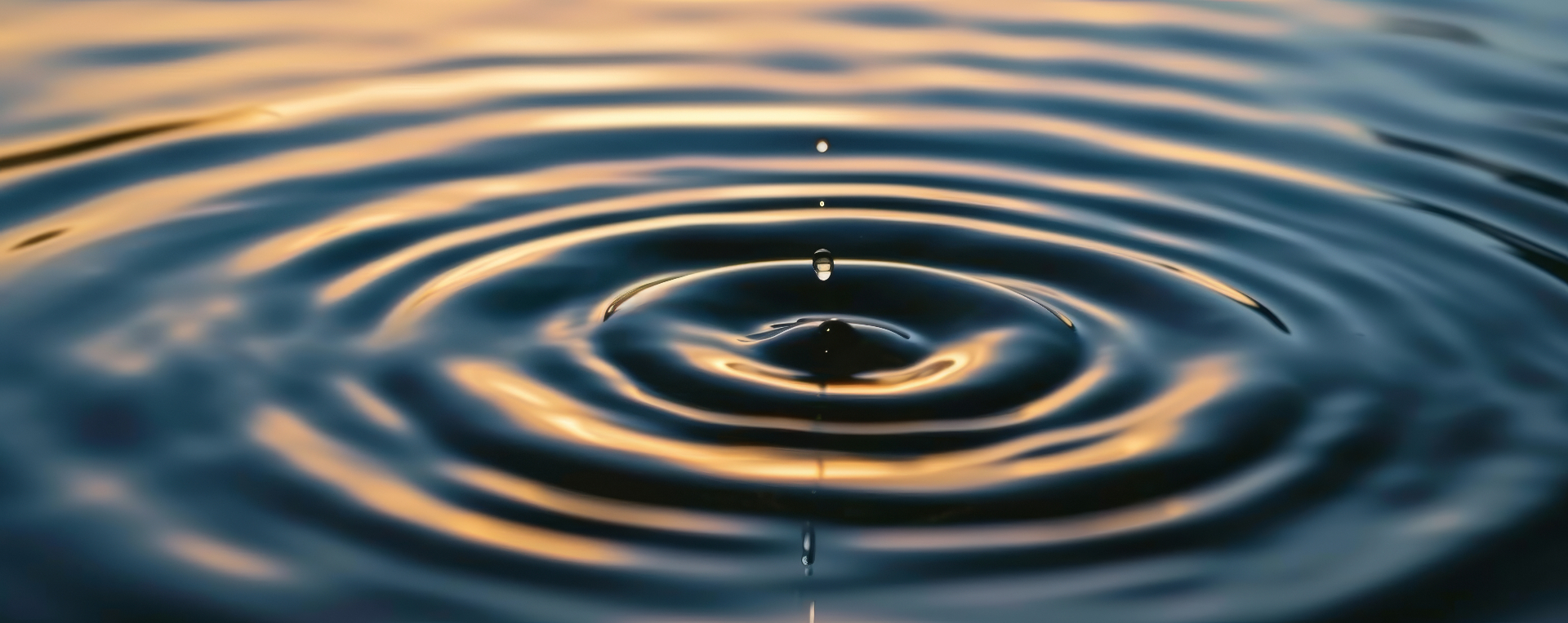 A single water droplet meeting a still, deep blue surface, sending calm ripples outward — a visual metaphor for small changes creating lasting momentum.